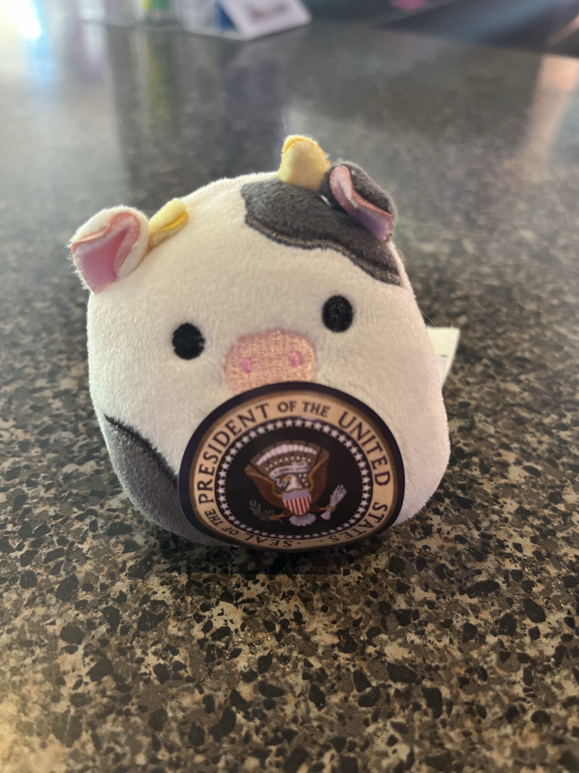 A tiny squishmallow cow, wearing a sticker with the United States Presidential seal